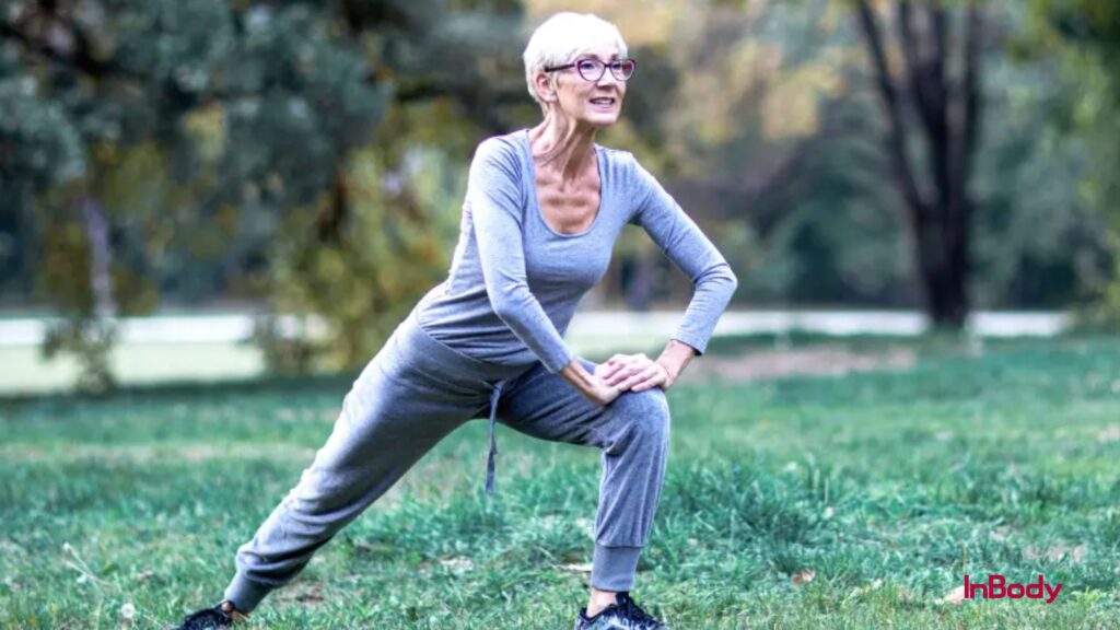 The image features an individual performing a stretching exercise outdoors in a park-like setting. They are dressed in a grey long-sleeve top and grey pants, positioned with one leg extended to the side while bending the other knee, with both hands resting on the bent knee. The surroundings include lush green grass, trees, and a pathway, creating a peaceful natural atmosphere. This scene highlights the importance of flexibility and fitness, especially when exercised outdoors, where the environment can enhance the experience.
