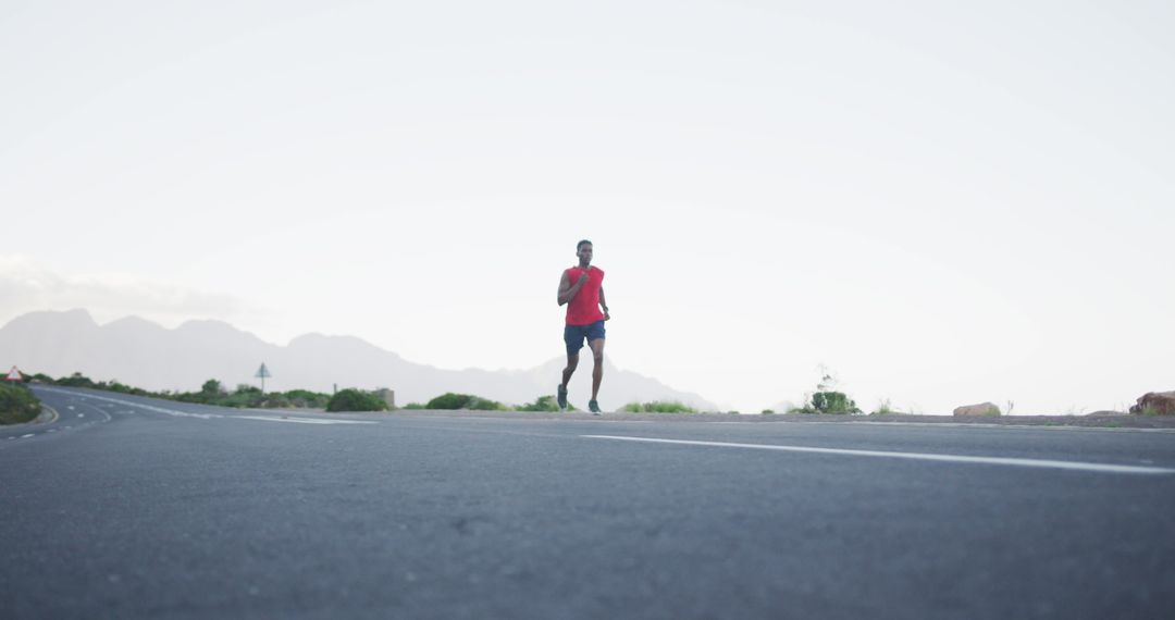 A man jogging alone on an open road with mountains in the background, wearing a red sleeveless shirt and blue shorts, symbolizing health and endurance.